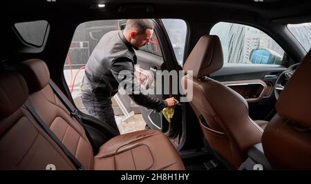 Blick von innen auf einen jungen Mann, der ein Innenauto auf der Autowaschanlage im Freien wascht. Schöner Arbeiter Polieren Automobil, mit Mikrofaser. Stockfoto