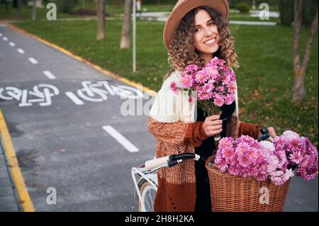 Attraktive junge Frau im Hut mit Blumenstrauß und lächelnd, während sie im Park mit dem Fahrrad unterwegs ist. Stilvolle Frau Fahrrad mit Blumen im Korb auf der Straße reiten. Stockfoto