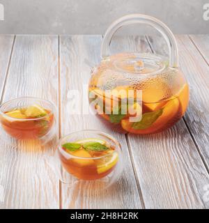 Früchtetee mit Orange, Zitrone und Minze in Glas-Teekannen und zwei Tassen (Schalen) auf Holzboden Stockfoto