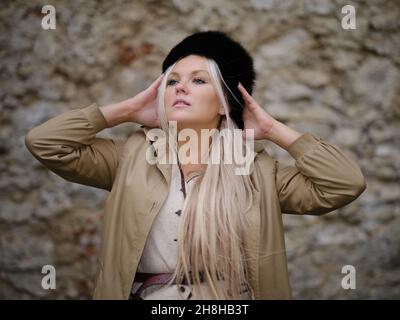 Blonde Frau mit schwarzem Pelzhut und beigem Mantel, die sich vor einer alten Steinmauer posiert, mit weichem natürlichem Licht und Wintermodeatmosphäre. Stockfoto