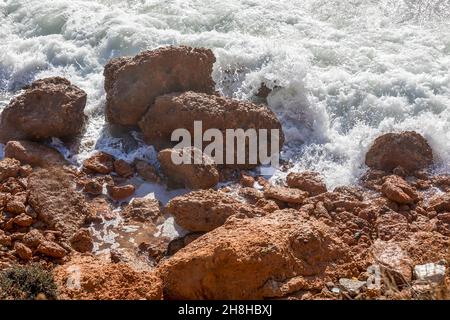 Seafoam. Meereswellen mit Schaum, die auf roten Felsen an der Küste des Kokina Beach auf der Insel Syros in Griechenland krachen. Stockbild. Stockfoto