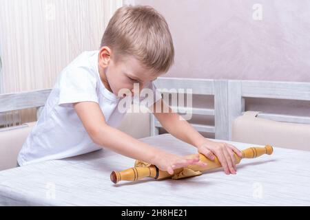 Das Kind rollt den Teig mit einem Nudelholz auf dem Tisch in der Küche aus. Der kleine Junge hilft der Mutter beim Kochen und macht selbstgemachte Gurkensaugen. Der Junge lernt Stockfoto