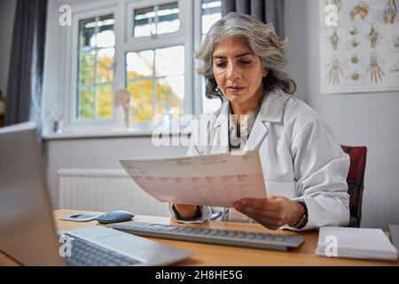 Ältere Frau GP trägt weißen Mantel am Schreibtisch in Arztpraxis Blick auf EKG-Anzeige Stockfoto