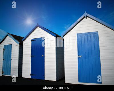 Drei blaue und weiße Strandhütten vor einem tiefen blauen Himmel mit der Sonne und dem hellen Strahl über dem Strand. Stockfoto