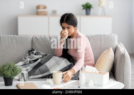 Verärgert traurige tausendjährige indische Frau mit Karli, an Grippe erkrankt, Kälte sitzt auf dem Sofa, Nase im Wohnzimmer blasen Stockfoto