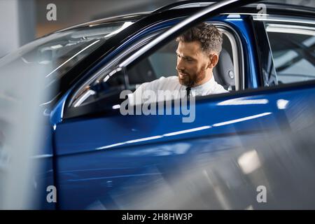 Kaukasischer bärtiger Mann in formellen Verschleiß testen Luxus-Auto im Auto Salon. Männlicher Kunde sitzt auf dem Fahrersitz mit offener Tür und berührender Lederausstattung. Stockfoto