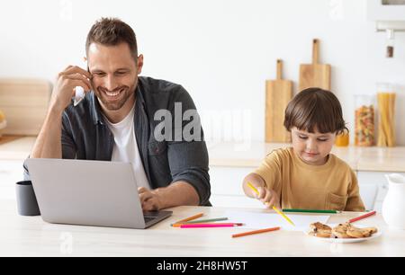 Netter kleiner Junge, der mit bunten Stiften Bilder zeichnet, in der Küche neben dem Laptop und dem Handy des Vaters sitzt Stockfoto