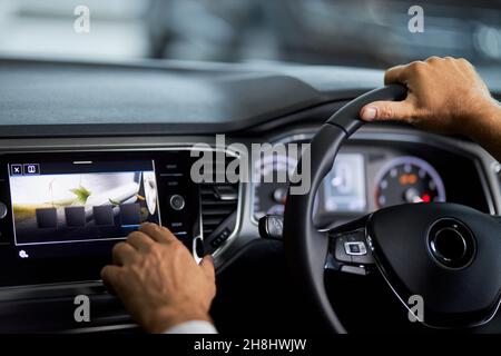 Mann, der in einem modernen Luxusauto sitzt und das digitale Display des Armaturenbretts berührt. Männlicher Kunde, der den Innenraum untersucht, während er sich für ein neues Fahrzeug im Ausstellungsraum entscheidet. Stockfoto