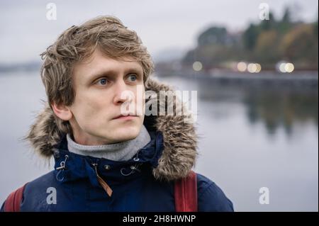 Porträt eines jungen, gutaussehenden Mannes mit blauen Augen in blauem Mantel an bewölktem Tag. Wasser mit Reflexion im Hintergrund. Stockfoto