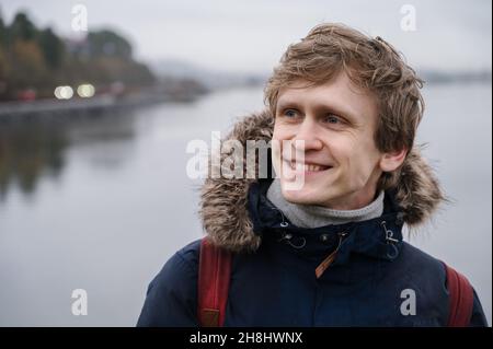 Porträt eines jungen, gut aussehenden lächelnden Mannes mit blauen Augen im blauen Mantel an bewölktem Tag. Wasser mit Reflexion im Hintergrund. Stockfoto