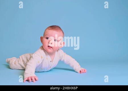 Der kleine Junge sieht neugierig aus, während er auf seinem blauen Hintergrund auf dem Bauch liegt. Vier Monate altes Kind, Speicherplatz kopieren Stockfoto