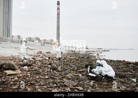 Weitwinkelaufnahme von Arbeitern, die in Gefahrgutanzügen arbeiten, die Proben der Sonde mit Wasser im industriellen Bereich sammeln, Konzept für giftige Abfälle, Kopierraum Stockfoto