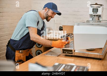 Fröhlicher junger Mann, der die Kaffeemaschine im Café repariert Stockfoto