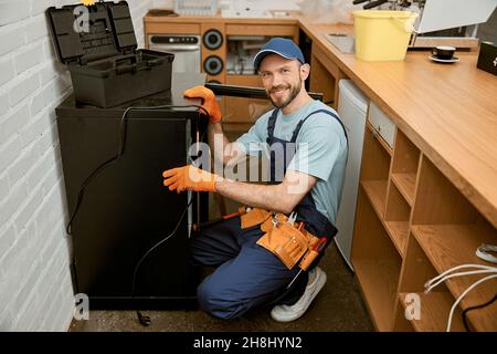 Fröhlicher junger Mann, der Kühlschrank in der Küche repariert Stockfoto