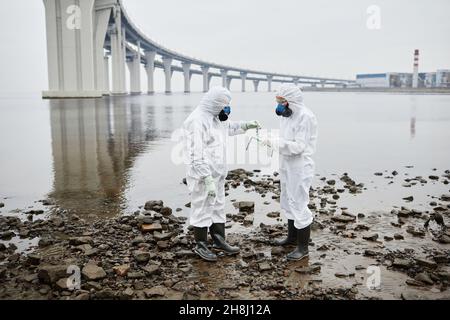 In voller Länge Porträt von zwei Personen in einem Hazmat-Anzug, der neben Wasser steht, giftiger Abfall und Schadstoffemissionen, Kopierraum Stockfoto