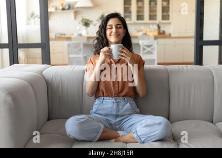 Hübsche junge kaukasische Dame, die mit gekreuzten Beinen auf der Couch sitzt und zu Hause frischen aromatischen Kaffee mit geschlossenen Augen riecht Stockfoto