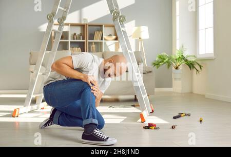 Der junge Mann, der gerade von der Leiter gefallen ist, sitzt auf dem Boden und umarmt sein verletztes Knie Stockfoto