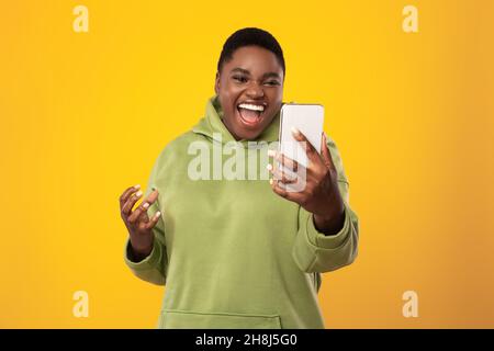 African American Lady Holding Smartphone Brüllt Emotional Über Gelben Hintergrund Stockfoto