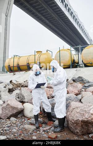 In voller Länge Porträt von zwei Wissenschaftlern in Gefahrgutanzügen, die Proben im Freien, toxische Abfälle und Verschmutzungskonzept sammeln Stockfoto