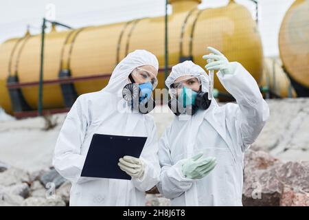 Waist-up-Porträt von zwei Wissenschaftlerinnen in Hazmat-Anzügen, die Proben im Freien sammeln, toxischen Abfall und Verschmutzungskonzept Stockfoto