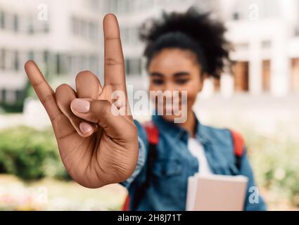 Hübsche, tausendjährige Studentin, die im Freien auf dem College-Campus große Felsgestikte zeigt. Teenager-Musikkonzept Stockfoto