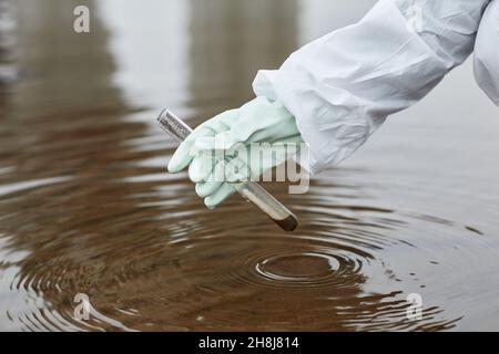 Nahaufnahme eines nicht erkennbaren Wissenschaftlers, der einen Hazmat-Anzug trägt, um Wasserproben zu sammeln, Fokus auf Handschuhen, die das Reagenzglas halten, Kopierraum Stockfoto