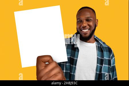 Glücklicher junger schwarzer Kerl, der ein weißes Papierbanner mit Platz für Werbung zeigt und Ihren Service auf orangefarbenem Hintergrund empfiehlt Stockfoto