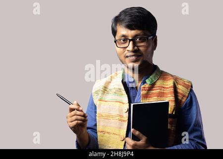 Ein junger indischer Mann in ethnischer Kleidung, der mit einem Buch steht und mit einem Stift auf grauem Hintergrund zeigt Stockfoto