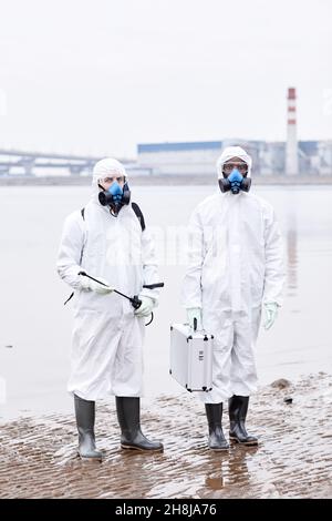 Vertikale Ganzkörperportrait von zwei Personen, die Schutzanzüge tragen und die Kamera auf einem Industriegelände im Freien betrachten Stockfoto