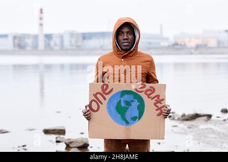 Waist-up-Porträt eines afroamerikanischen Mannes, der ein Erdzeichen in der Hand hält und die Kamera anschaut, während er am Standort der Umweltkatastrophe protestiert und den Weltraum kopiert Stockfoto