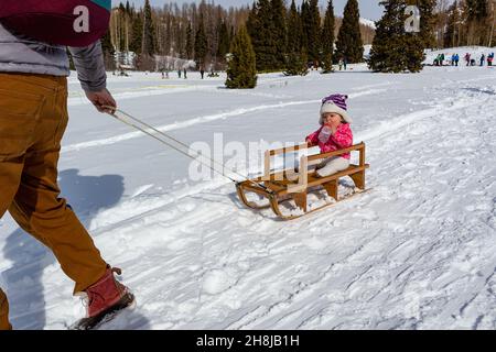 Ein kleines Mädchen, das aus einer Babyflasche trinkt, sitzt auf einem Holzschlitten, der von ihrem Vater während des Chama Chile Ski Classic gezogen wird. Stockfoto