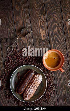 Draufsicht auf zwei französische Eclare und eine Tasse Espresso auf einem Holztisch gegen eine Streuung von Kaffeebohnen, Draufsicht mit Kopierraum Stockfoto