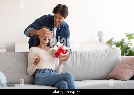 Lächelnder junger europäischer Ehemann schließt die Augen vor seiner Frau und schenkt im Wohnzimmer eine Geschenkbox Stockfoto