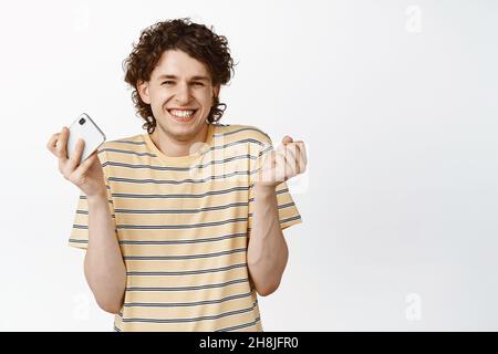 Enthusiastischer lockiger Kerl mit Mobiltelefon und Jubel, lächelnd aufgeregt, im T-Shirt auf weißem Hintergrund stehend Stockfoto