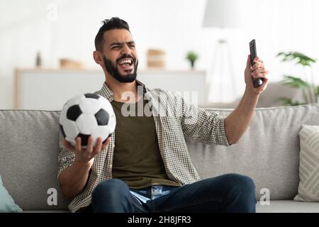 Fröhlicher junger Araber, der Fußballspiele im Fernsehen ansieht, Ball und Fernbedienung hält und zu Hause auf der Couch sitzt Stockfoto