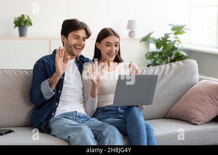 Ein fröhlicher, tausendjähriger europäischer Mann und eine fröhliche Dame sitzen auf der Couch, winken die Hände in die Webcam des Laptops und rufen online an Stockfoto