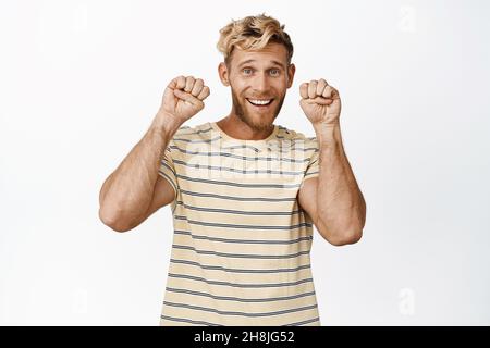 Maskuliner starker blonder Kerl macht niedliche Gesicht- und Handpfoten-Geste, lächelt glücklich an der Kamera, steht über weißem Hintergrund Stockfoto