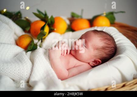 Kaukasisches Neugeborenes liegt in einem Korb mit Mandarinen unter einer weißen Decke. Stockfoto