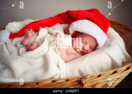 Das kaukasische schlafende Neugeborene in der Weihnachtsmütze des Weihnachtsmannes liegt in einem Korb unter einer weißen Decke, die Lichter brennen vor dem Hintergrund. Stockfoto