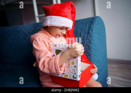 Ein kaukasisches Mädchen in einem rosa Frottee-Gewand und einer roten Weihnachtsmütze sitzt zu Hause auf der Couch und öffnet freudig ein Weihnachtsgeschenk, war das Mädchen überrascht. Stockfoto