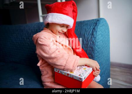 Ein kaukasisches Mädchen in einem rosa Frottee-Gewand und einer roten Weihnachtsmütze sitzt zu Hause auf der Couch und öffnet glücklich ein Weihnachtsgeschenk. Stockfoto