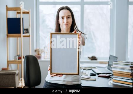 Rahmenbild, Plakat, Diplom, Urkunde in weiblichen Händen. Junge Brünette Frau hält leeren leeren hölzernen Fotorahmen auf Tisch mit Laptop Stockfoto