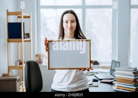 Rahmenbild, Plakat, Diplom, Urkunde in weiblichen Händen. Junge Brünette Frau hält leeren leeren hölzernen Fotorahmen auf Tisch mit Laptop Stockfoto