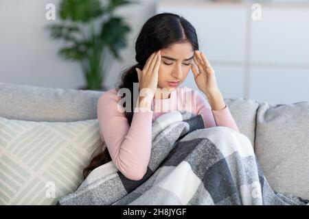 Traurige aufgeregte junge hindu-Dame, die mit einer Decke bedeckt ist, an Kopfschmerzen und Grippe leidet, sitzt auf dem Sofa und massiert die Schläfen Stockfoto