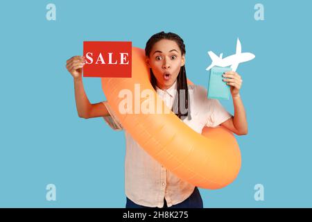 Porträt einer schockierten Frau mit Gummiring, mit Karte mit Verkaufsaufschrift und mit Erstaunen schauend, Sommerkäufe für Touren. Innenaufnahme des Studios isoliert auf blauem Hintergrund. Stockfoto