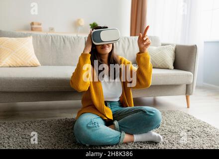 Fröhliche arabische junge Frau, die moderne VR-Brillen trägt, während sie im Wohnzimmer auf dem Boden sitzt und auf den Kopierraum zeigt Stockfoto