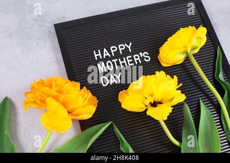 Schwarzer Karton mit weißen Plastikbuchstaben mit Zitat Happy Mothers Day und gelben Tulpen auf grauem Hintergrund. Stockfoto