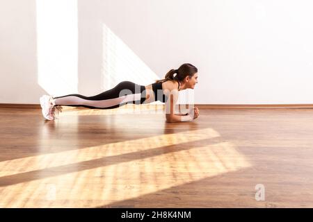 Seitenansicht einer sportlichen Frau, die Yoga praktiziert, Plank-Übungen an gebeugten Händen macht, Muskeln trainiert, schwarzes Sporttop und Strumpfhosen trägt. Studioaufnahme in voller Länge, die durch Sonnenlicht vom Fenster beleuchtet wird. Stockfoto