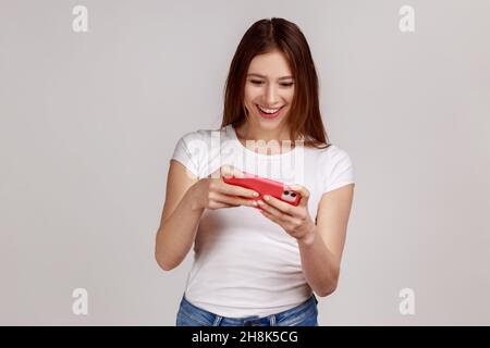 Liebenswert dunkelhaarige Frau mit Mobiltelefon mit aufmerksamen Gesichtsausdruck, Videospiel auf Handy spielen. Tragen weißes T-Shirt. Innenaufnahme des Studios isoliert auf grauem Hintergrund. Stockfoto