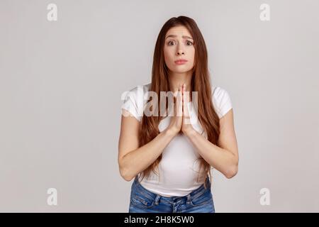 Bitte, das brauche ich. Junge Erwachsene Frau hält die Hände im Gebet und bittet um Hilfe bei flehenden, hoffnungsvollen Ausdrücken, die weißes T-Shirt tragen. Innenaufnahme des Studios isoliert auf grauem Hintergrund. Stockfoto